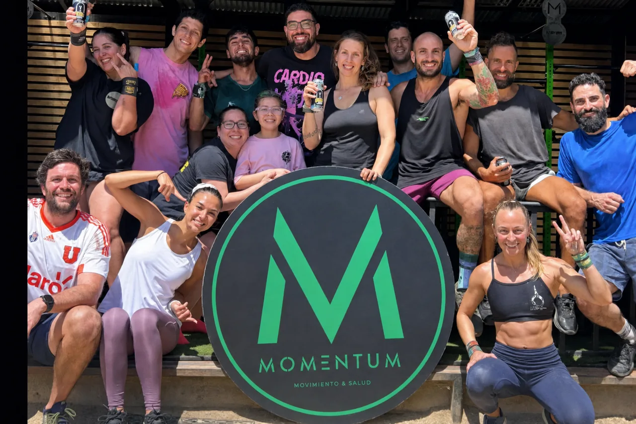 Un grupo grande de socios de Momentum, de edades y contexturas distintas, posando alrededor del logo físico después de una clase.